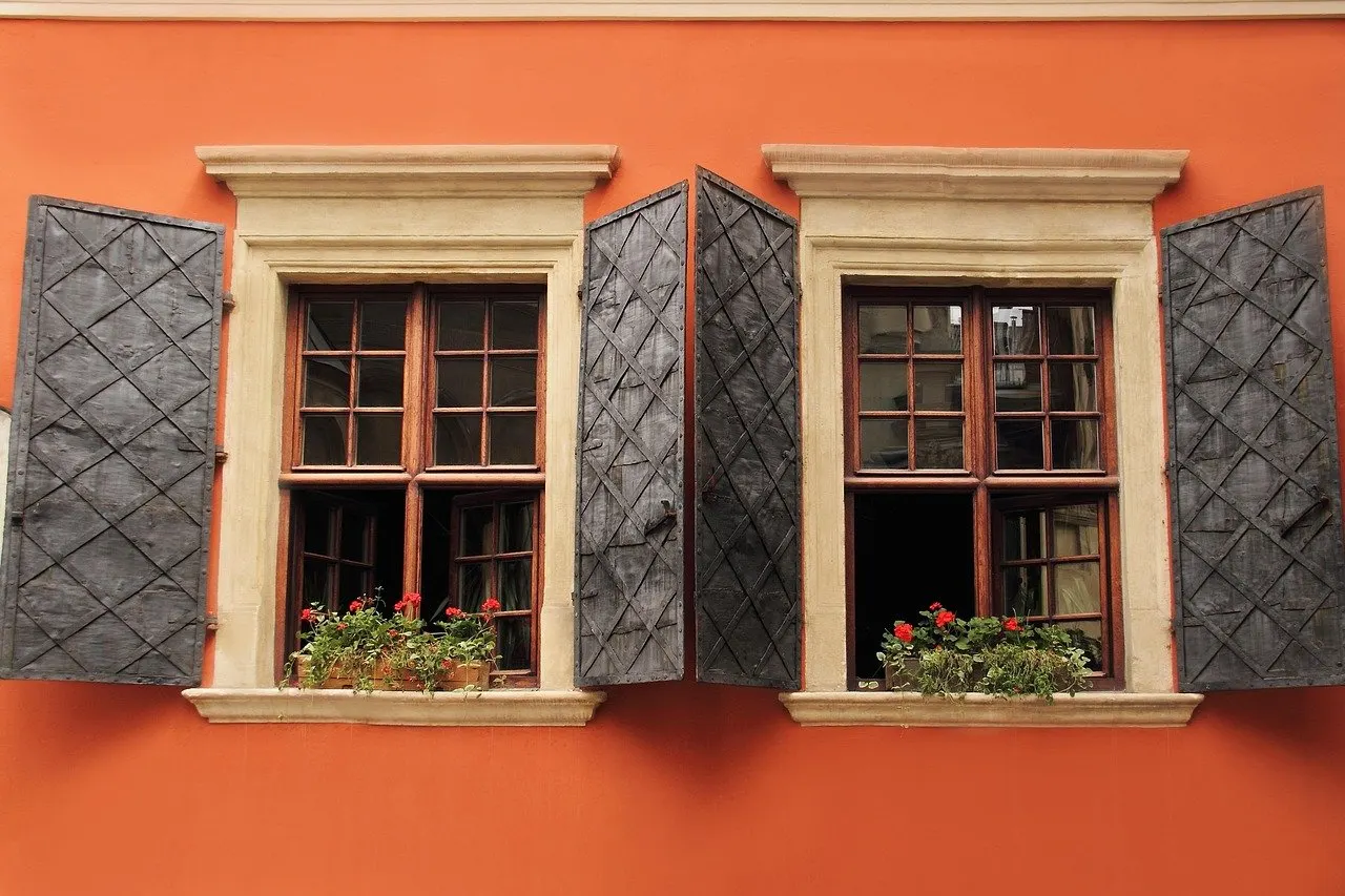Zwei Holzfenster mit Sprossen und offenen grauen Lamellenläden an orangefarbener Hauswand, Blumenkästen mit roten Blüten.